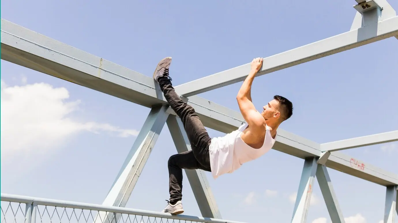 Side view of a young man climbing on the underside of a bridge.