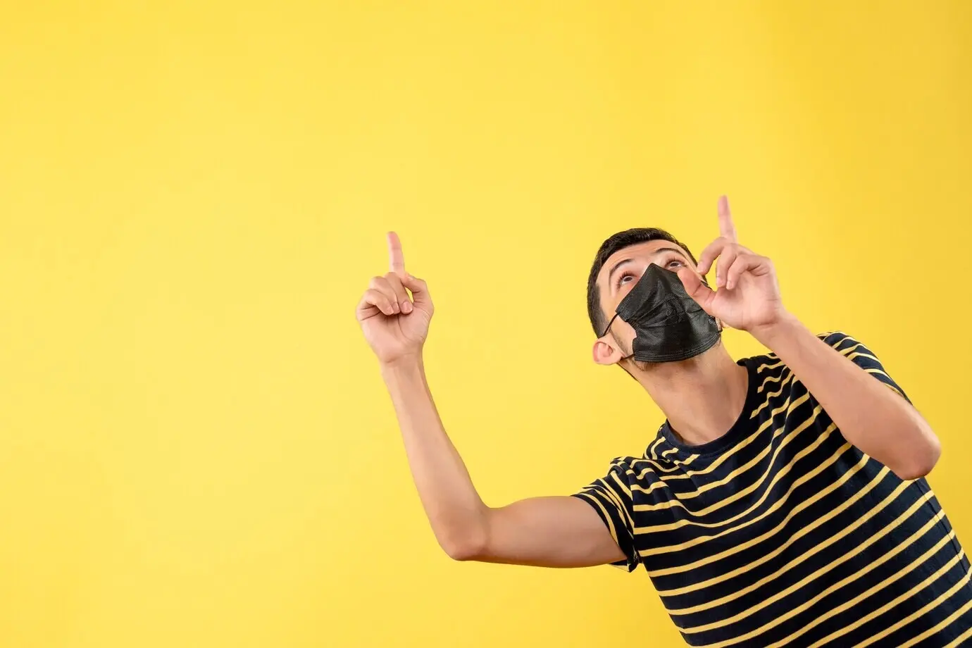 Frontal view of a handsome man in a black mask pointing at the ceiling against an isolated yellow background.