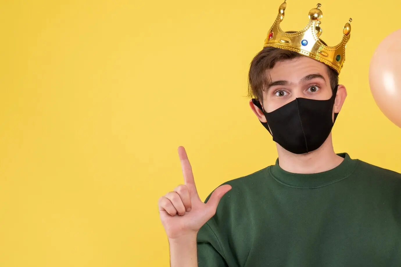 Front-facing young man wearing a black mask pointing at the ceiling on a yellow background.